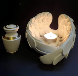 A lit candle sits in a white, wing-shaped holder beside a decorative urn on a dark background, evoking a serene and contemplative atmosphere.