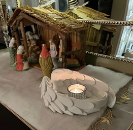 Nativity scene with figurines under a rustic stable roof on a decorative cloth. In the foreground, a lit candle is cradled in white angel wings.