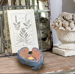 A cozy setup with a framed botanical print beside a lit tealight nestled in a blue, leaf-shaped holder. A stone-textured planter adds rustic charm.