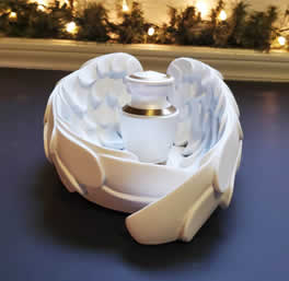 White angel wings candle holder cradles a small frosted urn with a metallic cap. Warm lights and greenery adorn the background, creating a serene atmosphere.