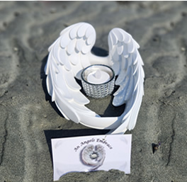 A white angel wing candle holder encircles a lit candle on sandy beach, conveying serenity. Below it, a card with an angel illustration reads An Angel's Embrace.