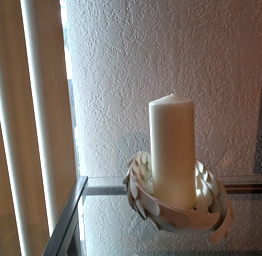 A white candle on a glass table is surrounded by a decorative holder, near vertical blinds and a textured wall, creating a calm, cozy ambiance.