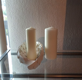 Two white pillar candles are placed on a glass shelf. One is surrounded by a delicate angel wing candle holder. The scene is calm and minimalist.