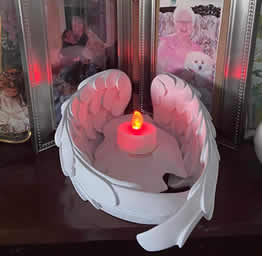 An angel wing candle holder with a glowing red-orange candle in the center is placed in front of framed family photos, creating a warm, sentimental ambiance.